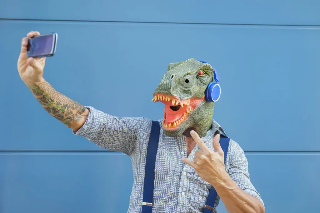 Crazy senior man wearing t rex mask while taking selfie with mobile smartphone Hipster male having fun listening music and dancing outdoor Absurd surreal and technology concept